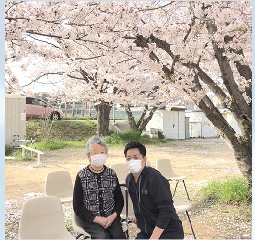 桜の下の椅子に腰かけるご利用者様と、そばに寄り添う理学療法士